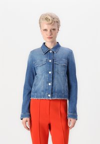 Jeansjacke mit einem hellen bis dunklen blauen Farbverlauf, zwei Brusttaschen, silbernen Knöpfen und einem rohen Saum. Kombiniert mit leuchtend roten Hosen.