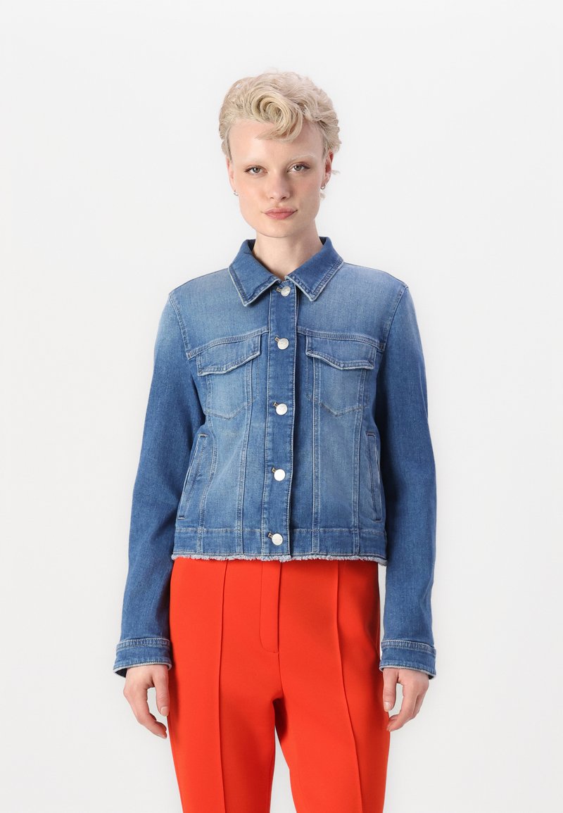 Jeansjacke mit einem hellen bis dunklen blauen Farbverlauf, zwei Brusttaschen, silbernen Knöpfen und einem rohen Saum. Kombiniert mit leuchtend roten Hosen.