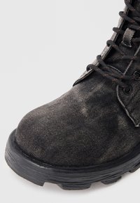 Bota de tobillo de mezclilla negra con punta redondeada, suela de goma y cordones negros. Presenta tela texturizada y detalles de costuras en contraste de color naranja.