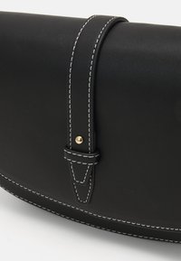 Mustasta nahasta valmistettu laukku, jossa on kaareva muoto, valkoiset ompeleet, kultainen koristehaka ja hihna, joka on kiinnitetty tyylikkäällä lenkillä.