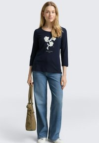 Marineblaue Langarmbluse mit weißem Floralgraphic und dem Text "ALPINE ESCAPE" darüber. Kombiniert mit hellblauen, weiter geschnittenen Jeans und einer beigefarbenen Schultertasche.