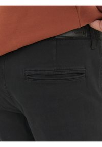 Pantaloni neri con una texture liscia, caratterizzati da un design della tasca posteriore cucita. Una felpa color ruggine è parzialmente visibile sopra.
