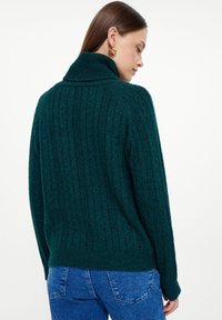 Maglione verde a maglia tricot con colletto alto e texture a coste. La vista posteriore mette in mostra la vestibilità comoda e il tessuto morbido.