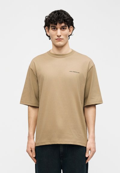 Νεαρός άνδρας με σγουρά σκούρα μαλλιά που φοράει ένα μπεζ φαρδύ T-shirt με μικρό μαύρο κείμενο στο στήθος, στέκεται μπροστά σε ένα απλό φόντο.