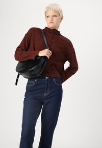 Sarkans teksturēts džemperis ar augstu apkakli, pārī ar tumši ziliem džinsiem. Melna plecu soma ar izliekta dizaina ir pāri plecam.