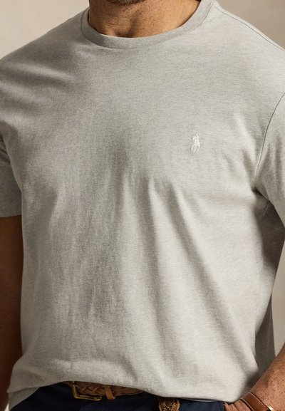 Άνδρας που φοράει ανοιχτογκρι μπλουζάκι με στρογγυλή λαιμόκοψη και μικρό λευκό κεντητό λογότυπο στο αριστερό στήθος, συνδυασμένο με καφέ πλεκτή ζώνη.