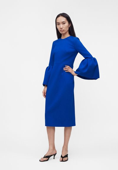 Γυναίκα στέκεται με το χέρι στη μέση, φοράει ηλεκτρικά μπλε midi φόρεμα με φαρδιά μακριά μανίκια και μαύρα σανδάλια με τακούνι σε λευκό φόντο.