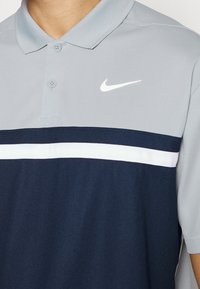 Polo tričko Nike s světle šedou horní částí a navy spodní polovinou s vodorovným bílým pruhem. Knoflíky u límce; měkká textilie.