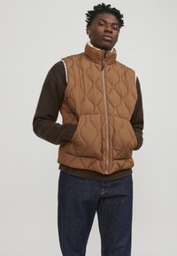 Quilted brown puffer vest with high collar, zip closure, and side pockets, layered over a dark brown sweater and blue jeans.