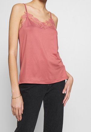 Frau, die ein schmutzig-pinkes Spaghettiträgertop mit Spitze trägt, kombiniert mit schwarzen Hosen, und vor einem schlichten Hintergrund steht.