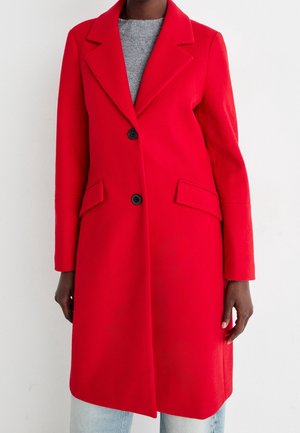 Femme portant un manteau rouge vif, mi-long jusqu'aux genoux, à simple boutonnage avec des boutons noirs et des poches à rabat, par-dessus un haut gris et un jean bleu clair.