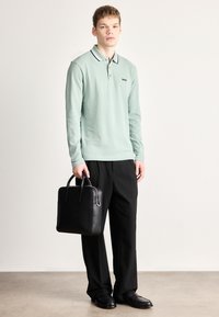 Polo de manga comprida verde claro com colarinho contrastante, logótipo no peito; calças pretas; a transportar uma pasta de couro preta; sapatos pretos.