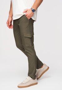 Homme portant un pantalon cargo vert olive et des baskets beige avec des lacets blancs, main dans la poche et montre-bracelet argentée au poignet gauche.