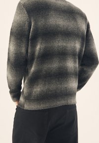 Pull en tricot aux tons dégradés de gris avec une texture douce, présentant un col rond et des poignets côtelés, associé à un pantalon noir.