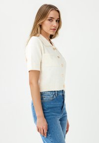 Kurzärmlige, cremefarbene Strickjacke mit kragenähnlichem Ausschnitt und Knopfverschluss, die eine weiche Textur und zwei vordere Taschen aufweist. Kombiniert mit blauen Jeans.