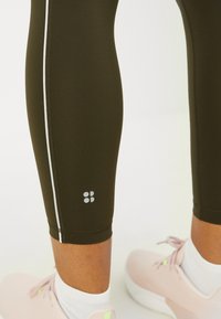Közelkép alsó lábakról, sötét olíva színű leggingsben, fehér oldalcsíkokkal, fehér zoknikkal és világos rózsaszín sportcipőkkel egy fehér háttér előtt.