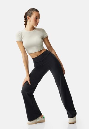 Giovane donna con capelli intrecciati che indossa un crop top bianco, pantaloni neri a gamba larga e sneakers bianche, piegata in avanti con le mani sulle ginocchia.