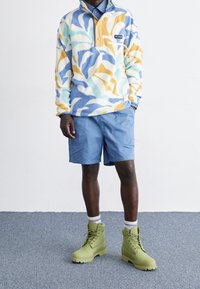 Pull en polaire bleu pastel, jaune et blanc avec un motif abstrait, doté d'un col zippé half; associé à un short bleu et des bottes vert olive.