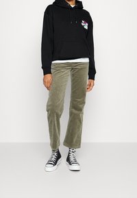 Sudadera con capucha negra y bolsillo frontal, combinada con pantalones de pana oliva y zapatillas altas negras con suelas y cordones blancos.