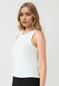Donna bionda con i capelli sciolti indossa un top bianco aderente senza maniche e pantaloni neri, in piedi contro uno sfondo chiaro uniforme.
