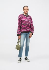 Pessoa em pé vestindo um suéter listrado rosa e castanho, jeans azul claro, sapatilhas pretas, segurando uma pequena mala verde.