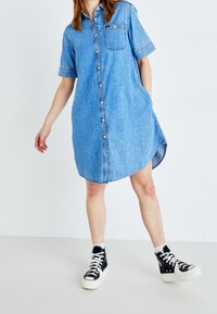 Abito in denim con maniche corte, chiusura a bottoni frontale, tasca sul petto e orlo curvo. Indossato con sneakers nere alte.