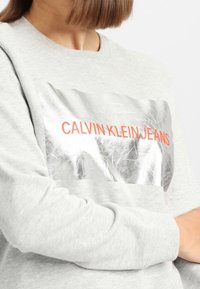 Sudadera gris con un panel plateado brillante que lleva "CALVIN KLEIN JEANS" en letras naranjas en negrita, cuello redondo y mangas largas.