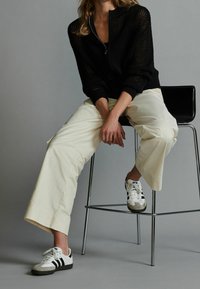 Schwarze gestrickte Zip-Up-Jacke, cremefarbene weite Hosen und weiße Sneakers mit schwarzen und grauen Akzenten, sitzend auf einem Metallhocker.
