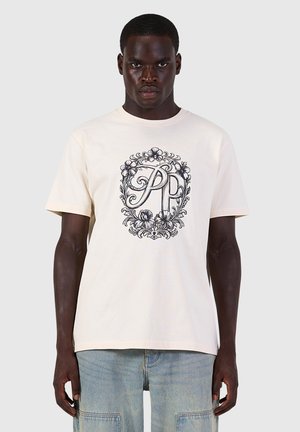 Man wearing a light beige T-shirt with a black ornate floral design and the letters P and P in the center, paired with light blue jeans.