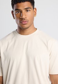 Vaaleanbeige puuvillapaita, jossa on pyöreä pääntie ja lyhyet hihat. Paidassa on sileä pinta ja rento istuvuus, ja se on päällä mallilla, joka katsoo suoraan eteenpäin.