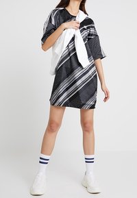 Mönstrad klänning i svart och vitt med ränder, prickar och geometriska former. Kort ärm och avslappnad passform, bärs med en vit tröja knuten runt halsen.