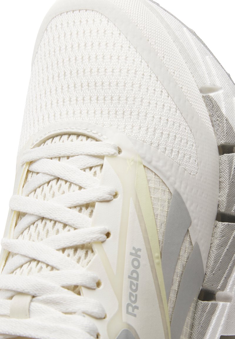 Zapatilla deportiva blanca con parte superior de malla, cordones planos y suela de goma con patrones texturizados y el logo de Reebok en el lateral.