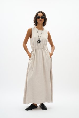 Nő ujjatlan bézs maxi ruhában oldalán megkötőkkel, fekete bebújós cipőben, fekete medálos nyaklánccal és fekete napszemüveggel, zsebében tartja a kezét.