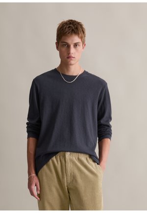 Langemouws marineblauw shirt van zacht materiaal met een ronde halslijn, gecombineerd met lichtgroene, geribbelde corduroybroek. Eenvoudige zilveren ketting als accent.