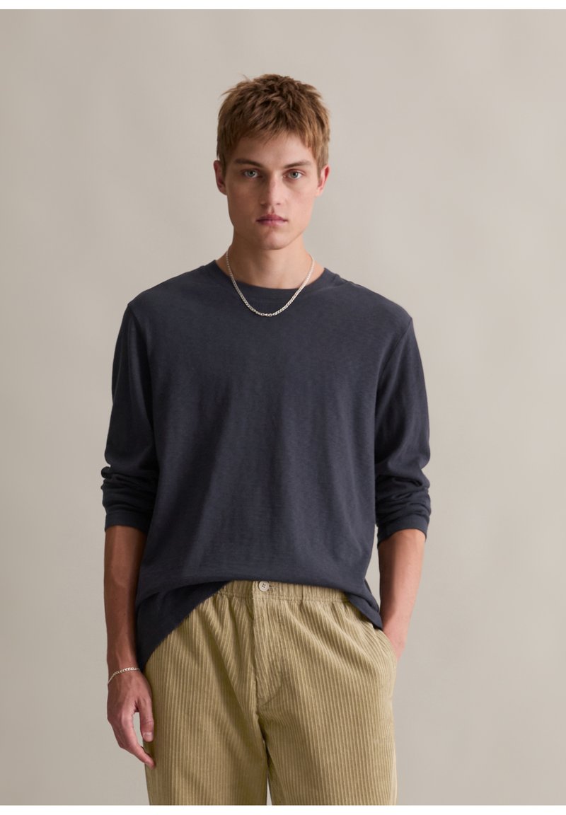 Langärmliges navy Hemd aus weichem Stoff mit Rundhalsausschnitt, kombiniert mit hellgrünen, gerippten Cordhosen. Einfache silberne Kettenakzent.