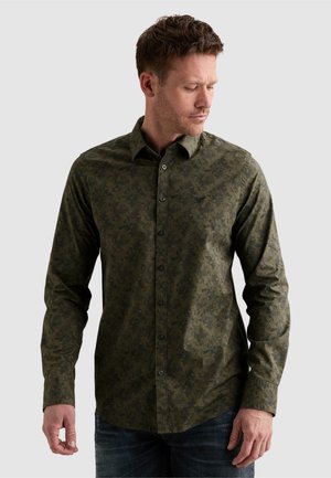 Man draagt een nauwsluitend donker groen patroon overhemd met lange mouwen en donkere jeans, kijkt naar links naar beneden tegen een effen achtergrond.