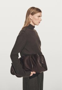 Brązowa skórzana torebka o miękkiej fakturze, z dwoma górnymi uchwytami i przeszyciami, zestawiona z ciemnym swetrem z dzianiny i szarymi spodniami.