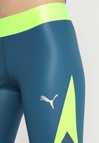 Leggings de tela suave y elástica en tono azul verdoso con una cinturilla amarilla brillante y un logotipo plateado destacado en el costado.
