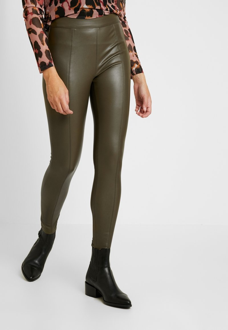 Olivgröna leggings i konstläder med hög midja, slät yta och sömnadsdetaljer, kombinerade med svarta ankelskor.