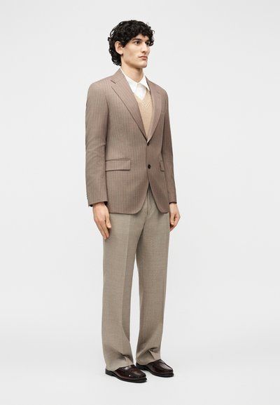 Άνδρας με σγουρά μαλλιά όρθιος, φοράει μπεζ σακάκι με λεπτές ρίγες, καφέ γιλέκο, λευκό πουκάμισο, φαρδιά μπεζ παντελόνια και σκούρα καφέ παπούτσια.