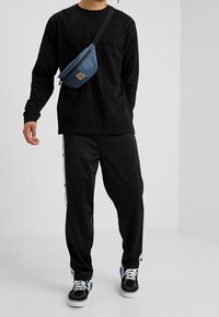 Langærmet sort skjorte, sorte bukser med sidelister, blå skuldertaske og sort-hvide sneakers. Enkel afslappet påklædning.