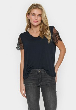 Top nero a maniche corte con scollatura a V e dettagli in pizzo sulle maniche, abbinato a jeans scuri. Il tessuto sembra liscio con una vestibilità rilassata.
