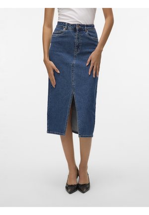 Kvinde iført en blå denim midi-nederdel med en frontslids, hvid top og sorte spidse hæle, stående mod en ensfarvet baggrund.