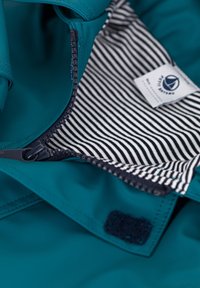 Veste teal avec une fermeture éclair à l'avant. L'intérieur est composé d'un tissu rayé et d'une étiquette de marque. Des matériaux texturés et un patch en Velcro sont visibles.