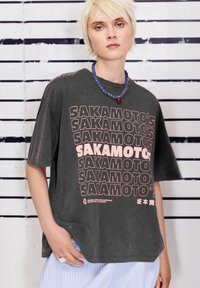 Šedé oversized tričko s opakujícími se růžovými nápisy "SAKAMOTO'S." Tkanina vypadá měkce, má volný střih a krátké rukávy.