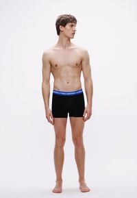 Μαύρα μποξεράκια με μπλε ζώνη μέσης που φέρει το λογότυπο "KARL LAGERFELD". Το μοντέλο στέκεται όρθιο, παρουσιάζοντας μια εφαρμοστή σχεδίαση.