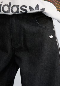 Jeans de ganga pretos com um pequeno logotipo prateado da Adidas no bolso frontal, complementados por riscas brancas nas laterais e um tecido texturizado.