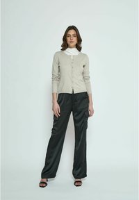 Cardigan beige con bottoni, abbinato a pantaloni larghi in satin neri con tasche e una consistenza liscia. Il modello è in piedi su uno sfondo neutro.