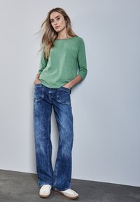 Vihreä pitkähihainen paita yhdistettynä sinisiin leveälahkeisiin farkkuihin. Paidassa on pyöreä pääntie ja farkuissa on etutaskut sekä kulutettu viimeistely.