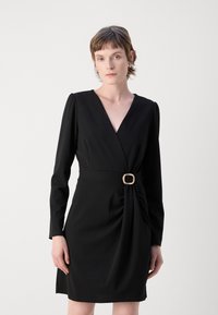 Robe noire portefeuille à manches longues, décolleté en V et détail de torsion sur le côté fixé par une boucle rectangulaire de couleur claire. Texture de tissu lisse.